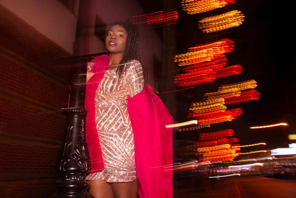 A black woman in sparkly dress is taking bright pink fur coat off her shoulder on a street at night