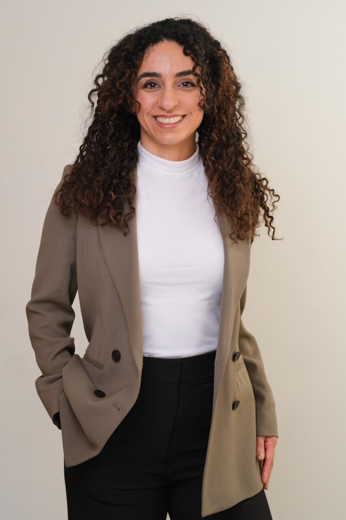 professional business portrait, woman with curly hair and big smile wearing a white top and a long grey blazer in front of white wall