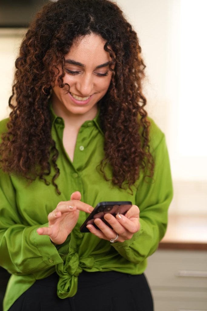 social media marketing content photoshoot with a freelance professional. Woman with dark curly hair wearing a green shirt smiling when typing on the phone